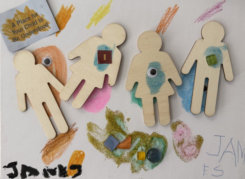 Mixed-media artwork on canvas featuring four wooden human figures decorated with googly eyes, gem stickers, and painted color bursts in orange, pink, teal, and yellow. The name "James" is written twice in the lower corners.