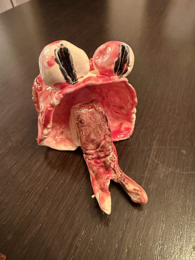 A small hand-built ceramic sculpture with bold red glaze sits on a dark wood surface. The figure has two round white eyes with heavy black markings and a hollow, arch-shaped body open at the front. A small limb extends from the base.